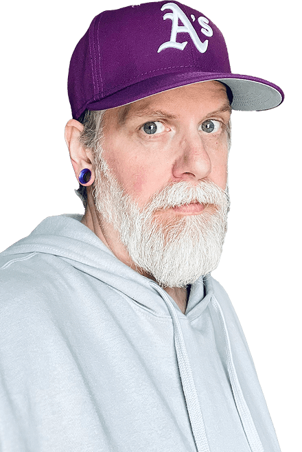 Photograph of James looking coyly at the camera, he’s wearing a light grey hoodie, a purple Oakland Athletics baseball cap and matching 1/2 inch gauge tunnels. His beard is well groomed and grey and white.